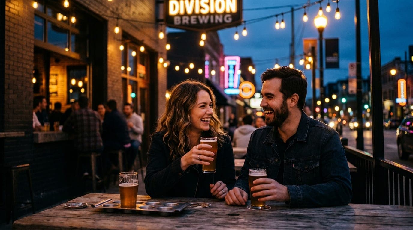 A couple laughs over craft beers at a rustic wooden table at Division Brewing, framed by warm string lights and deep shado...