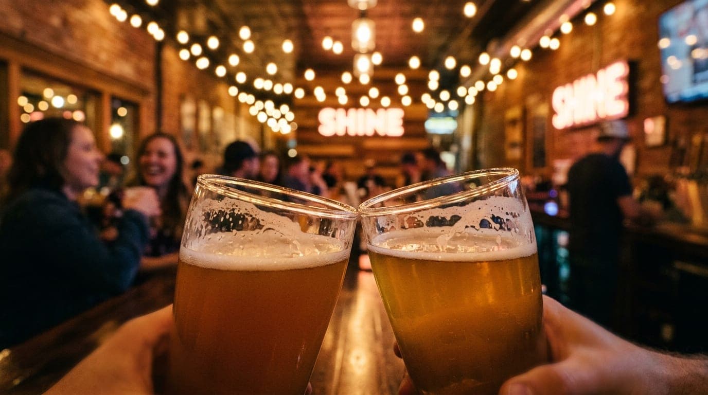 A tight 100mm macro shot of two craft beer glasses clinking against the warm, glowing backdrop of the Shine Beer Sanctuary...