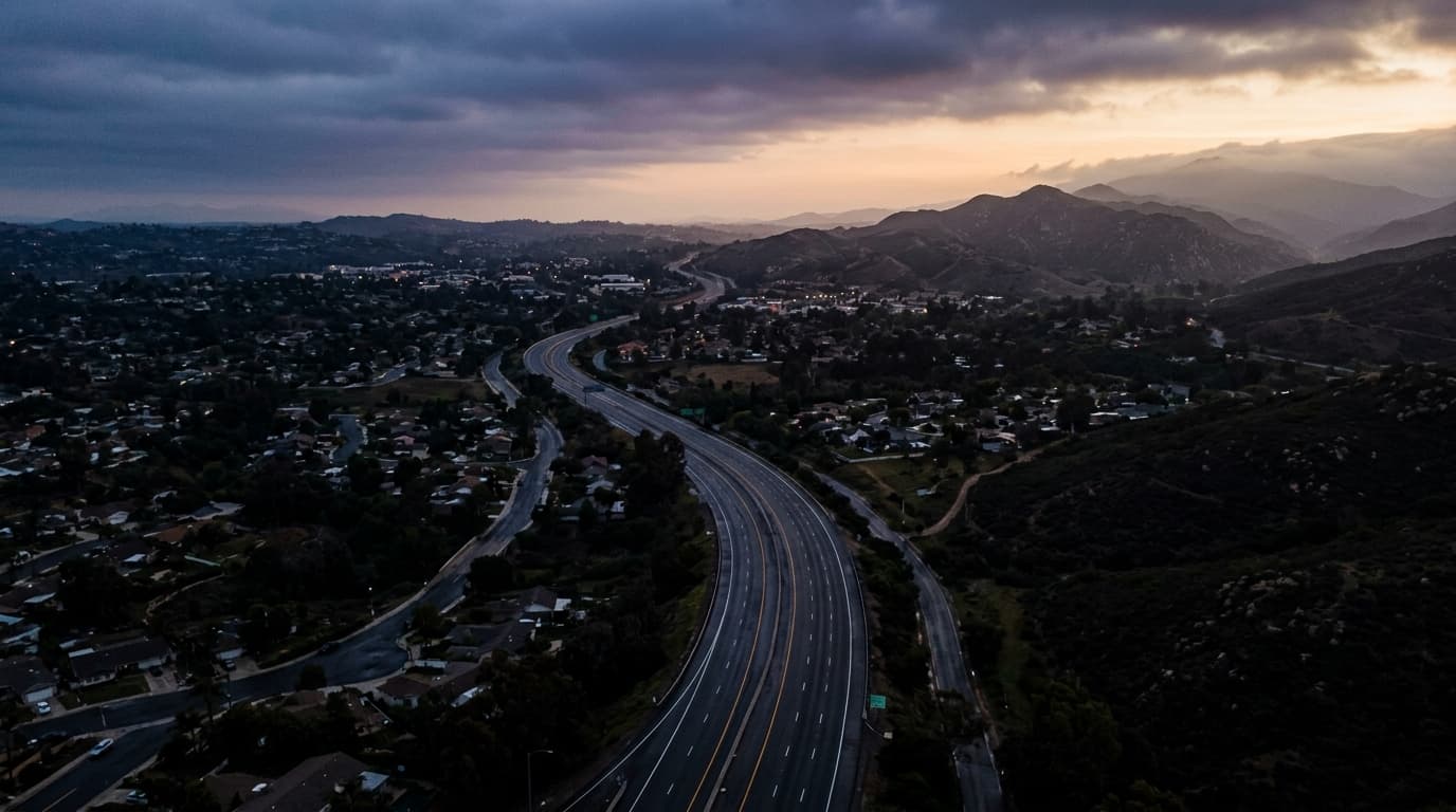 Overhead flat-lay 35mm shot of a winding, empty suburban highway ribboning through the darkened Escondido valley, captured...