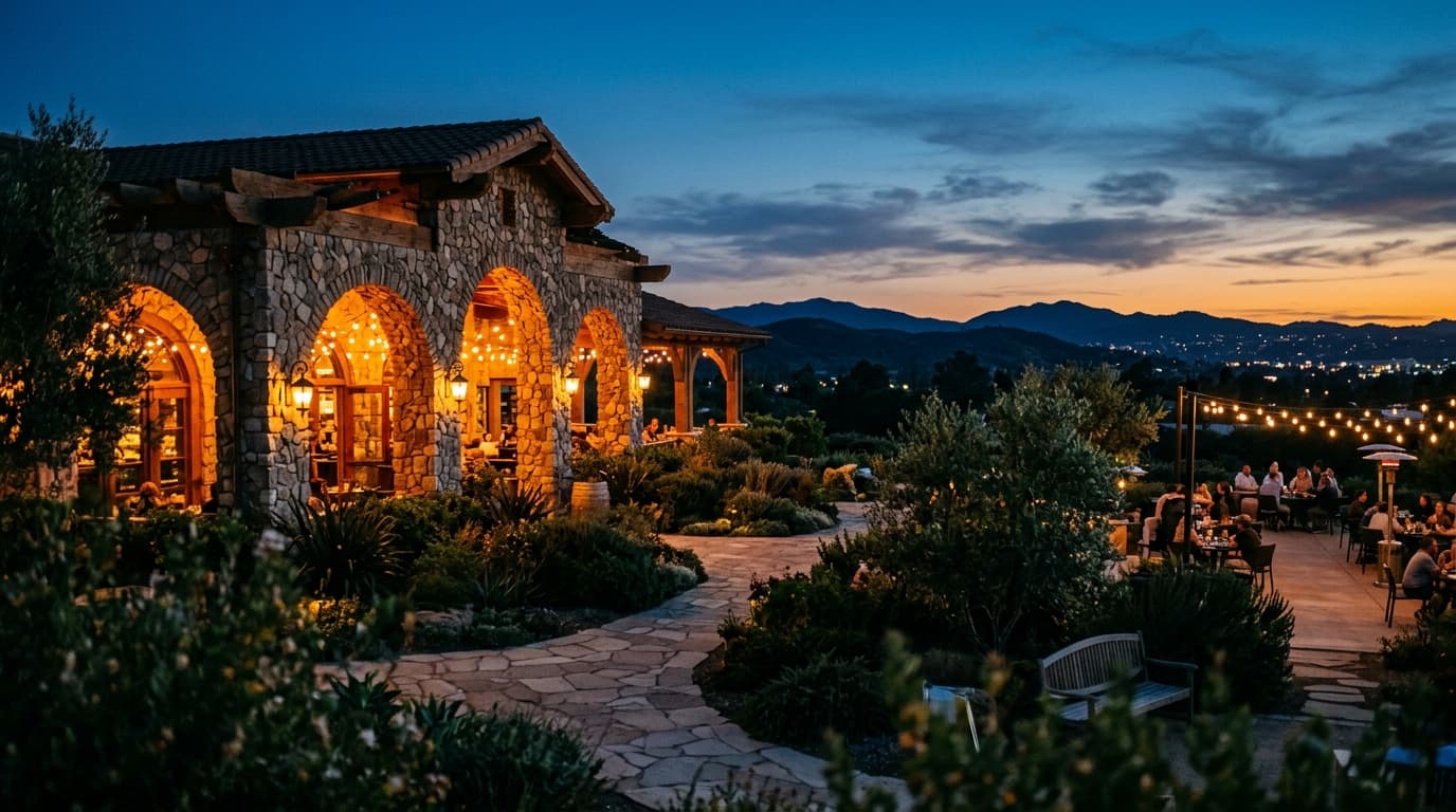 A wide 85mm shot of the Stone Brewing World Bistro gardens at dusk. Warm tungsten lights glow from the architectural stone...