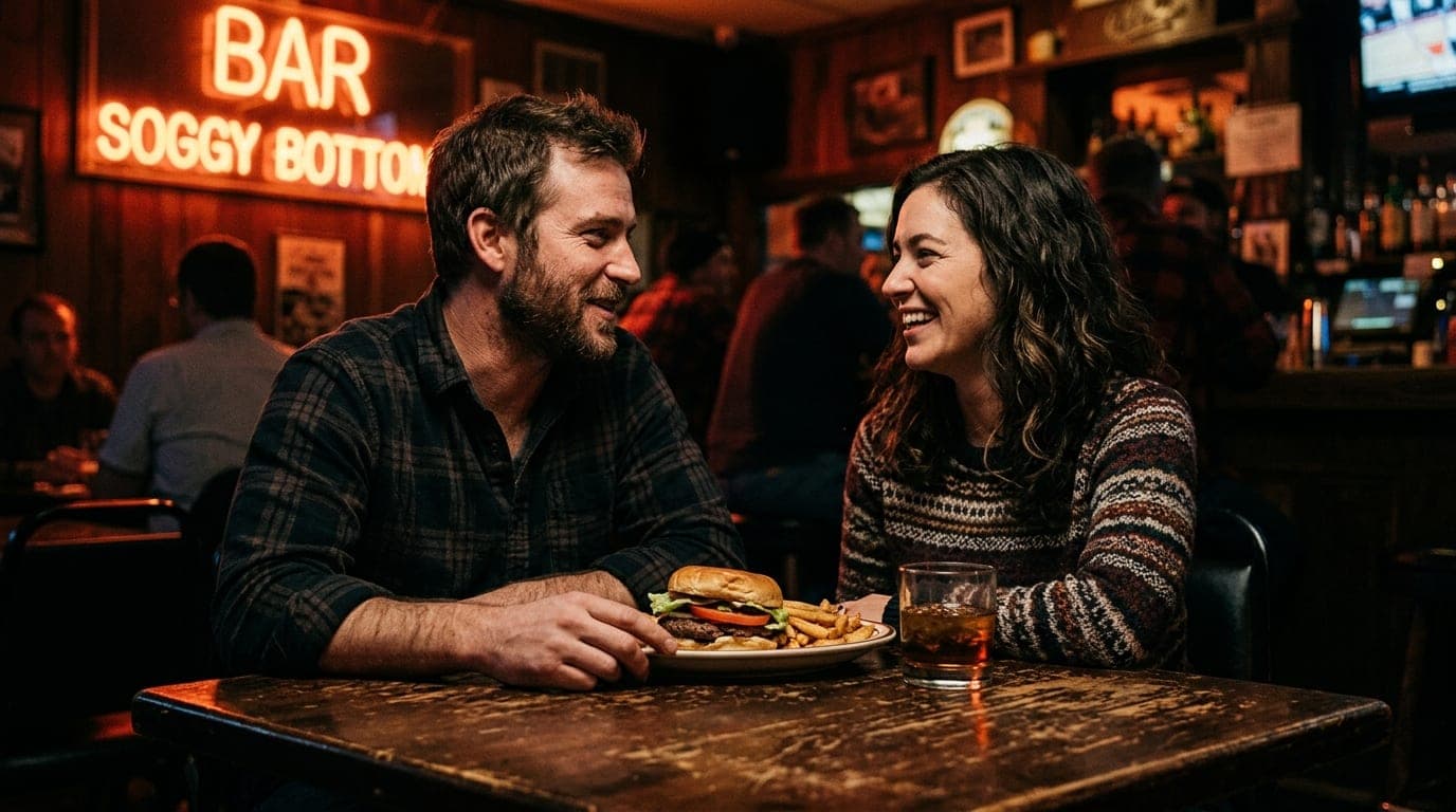 A couple sits at a scratched, dark wood table inside Soggy Bottom Bar. A half-eaten burger and a whiskey glass sit between...