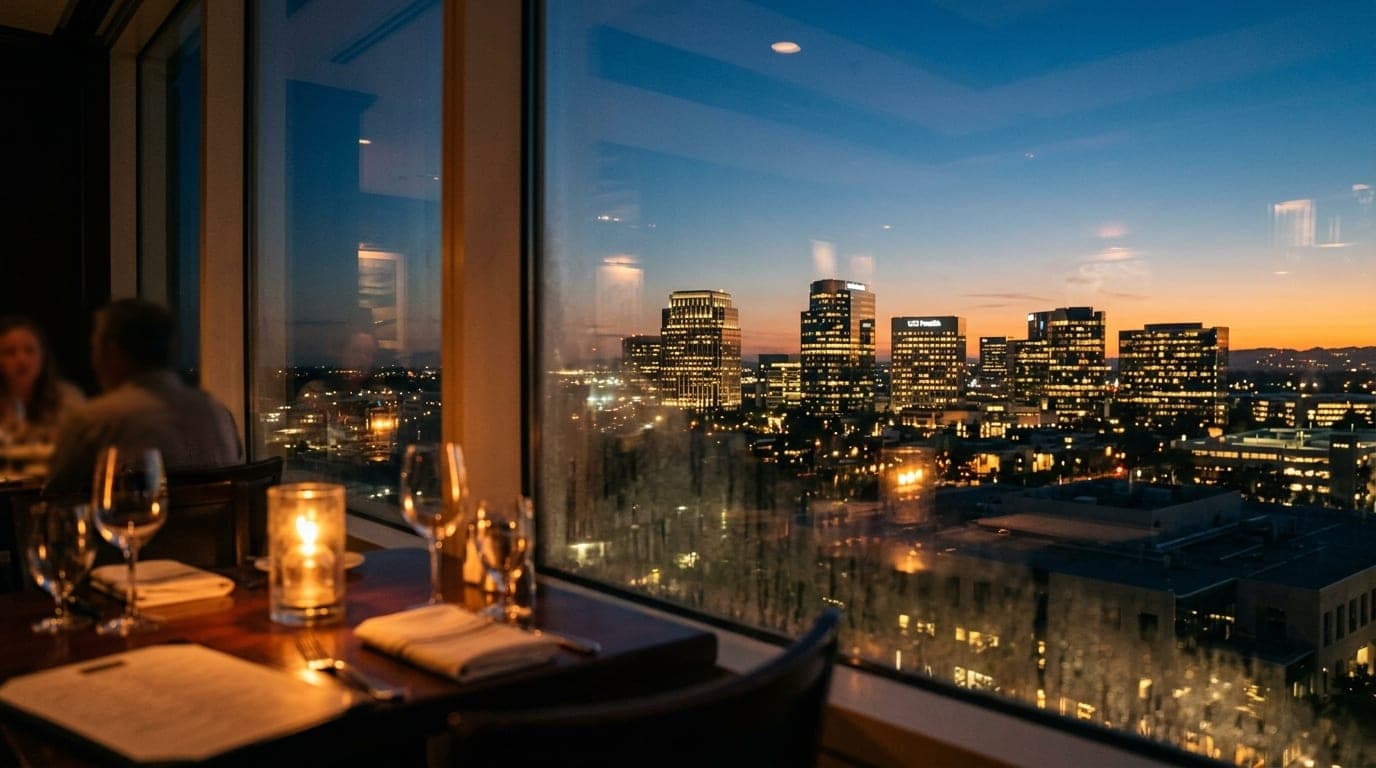 A soft, candle-lit table interior at Park Place overlooking the illuminated Irvine office towers at dusk, captured with an...