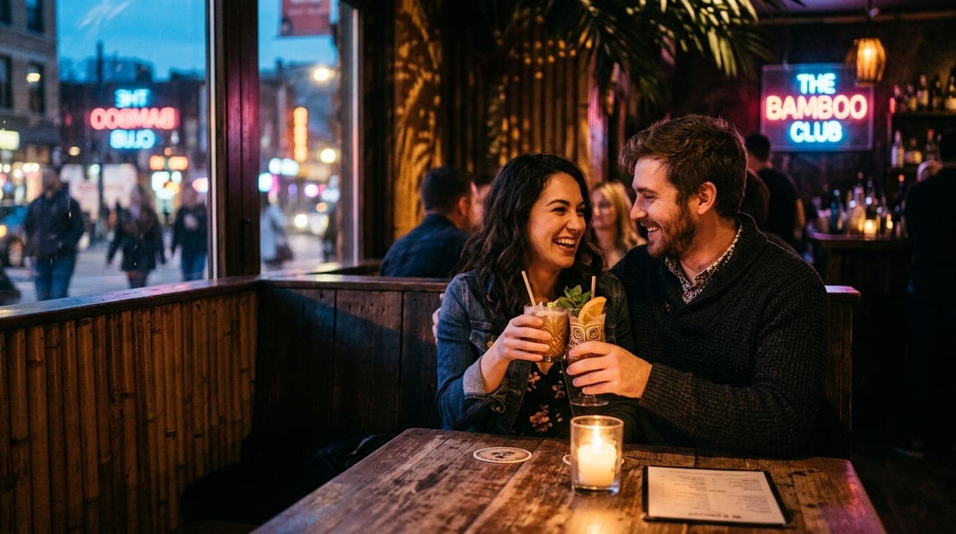A medium shot, offset right, captures a couple sharing a rum cocktail at a dark, candle-lit wooden booth in The Bamboo Clu...