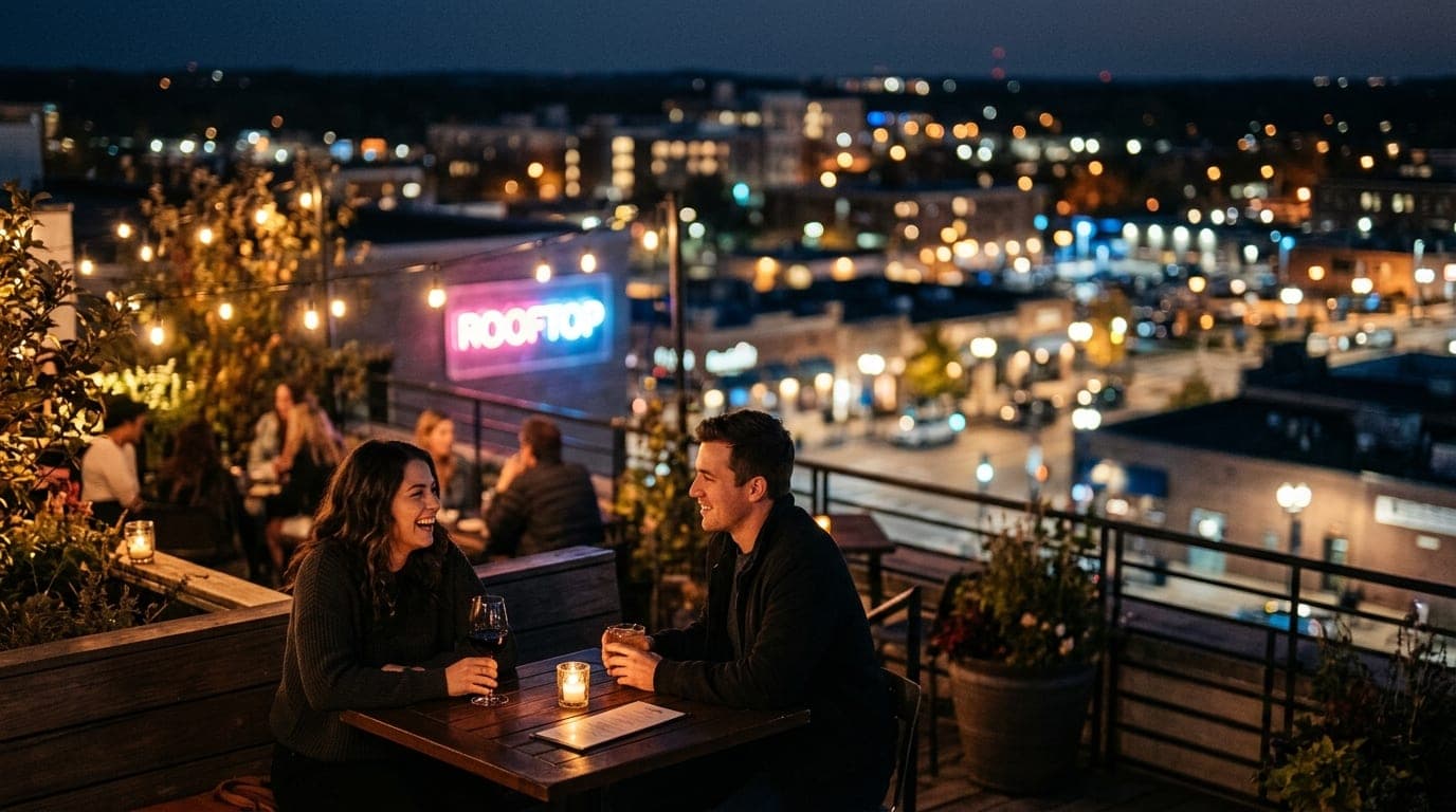 A couple sits at an outdoor corner table of a dimly lit rooftop bar, amber tungsten glow highlighting their faces. Behind...