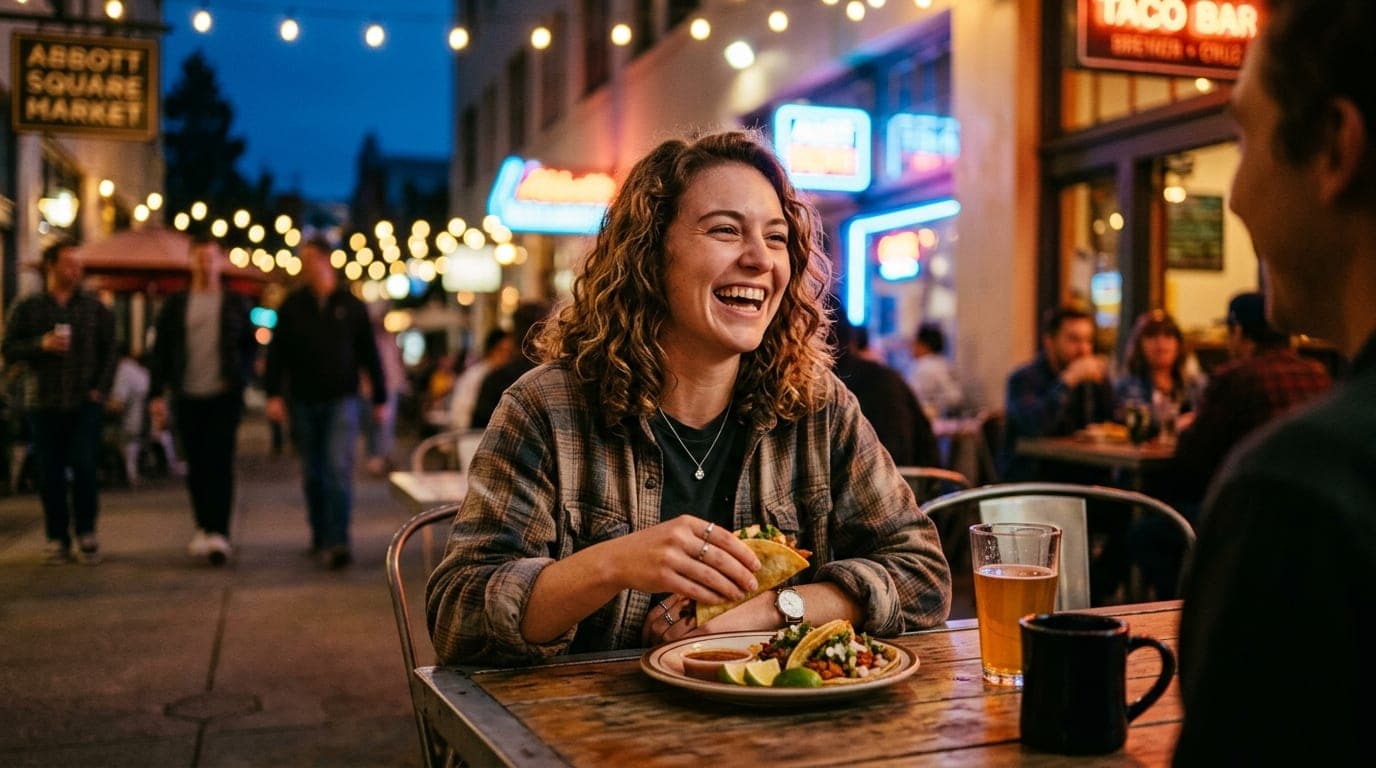 85mm portrait, shallow depth of field. A person in a casual flannel shirt sits at an outdoor Abbott Square table, laughing...