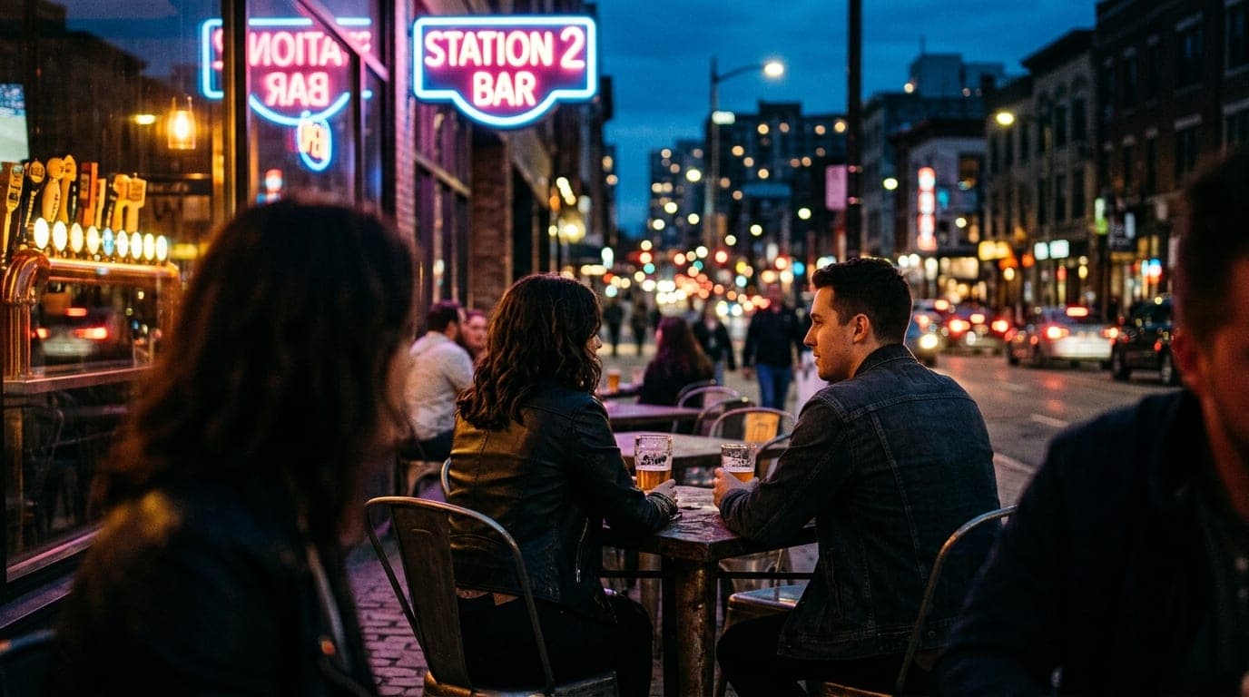 A couple in casual evening wear sits at a dark metal patio table at Station 2 Bar, seen over-the-shoulder, framed by glowi...