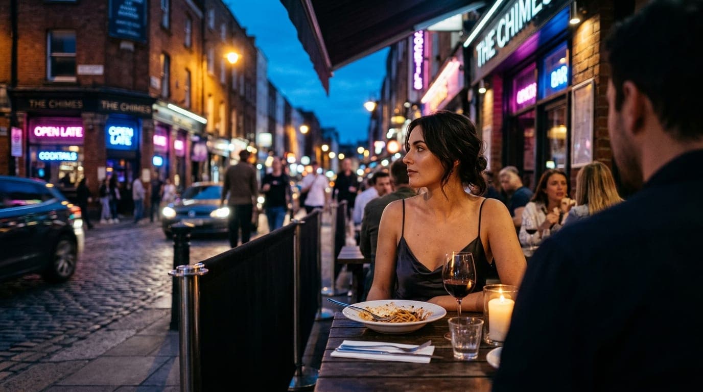 A woman in a silk slip dress sits at an outdoor table at The Chimes, a half-eaten late-night meal before her, backlit by b...