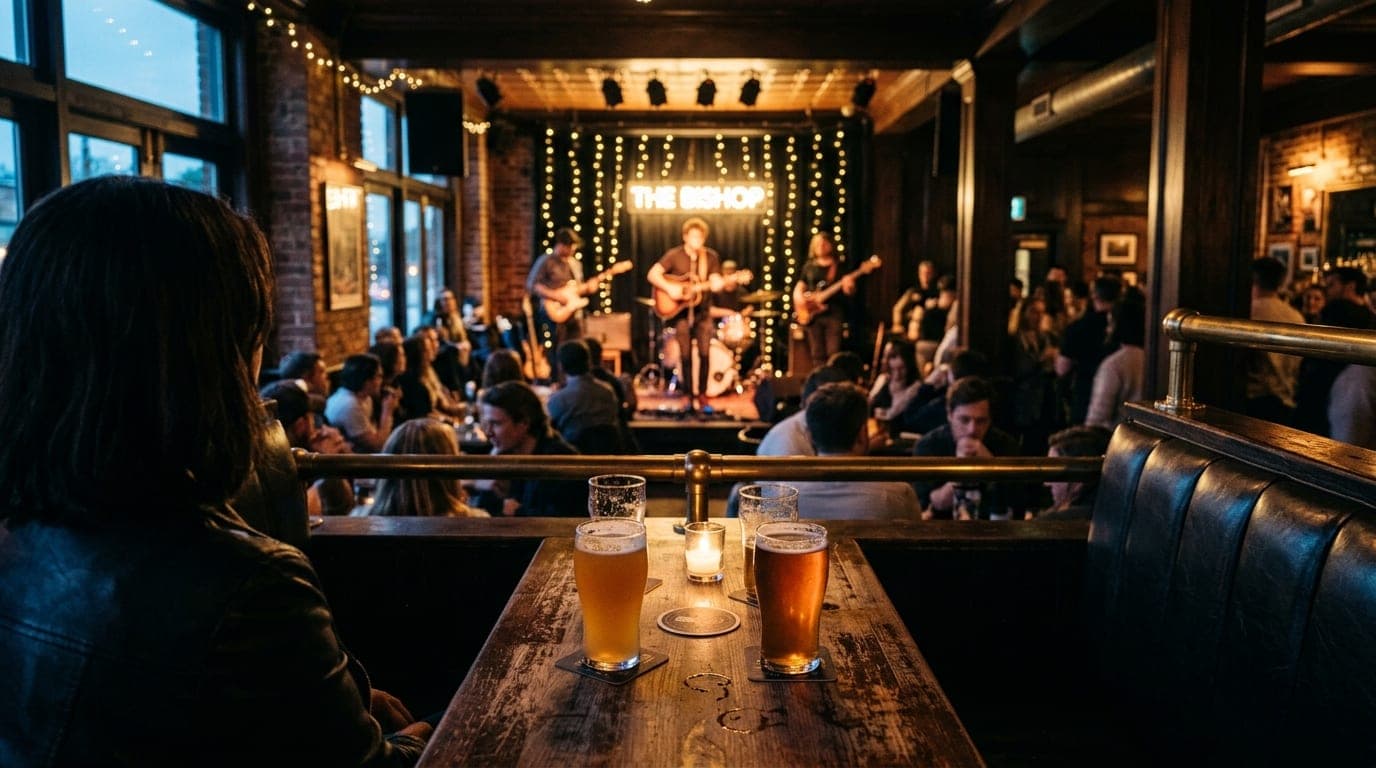 Over-the-shoulder view from a dark leather booth at The Bishop, looking toward the stage. Two cold craft beer pints sit on...