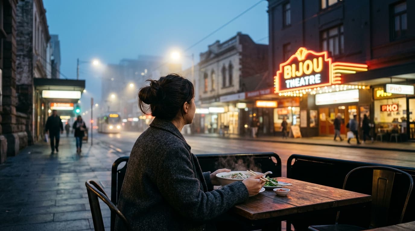 Over-the-shoulder shot at blue hour. A woman in a charcoal wool coat sits in the lower third, holding a steaming bowl of p...