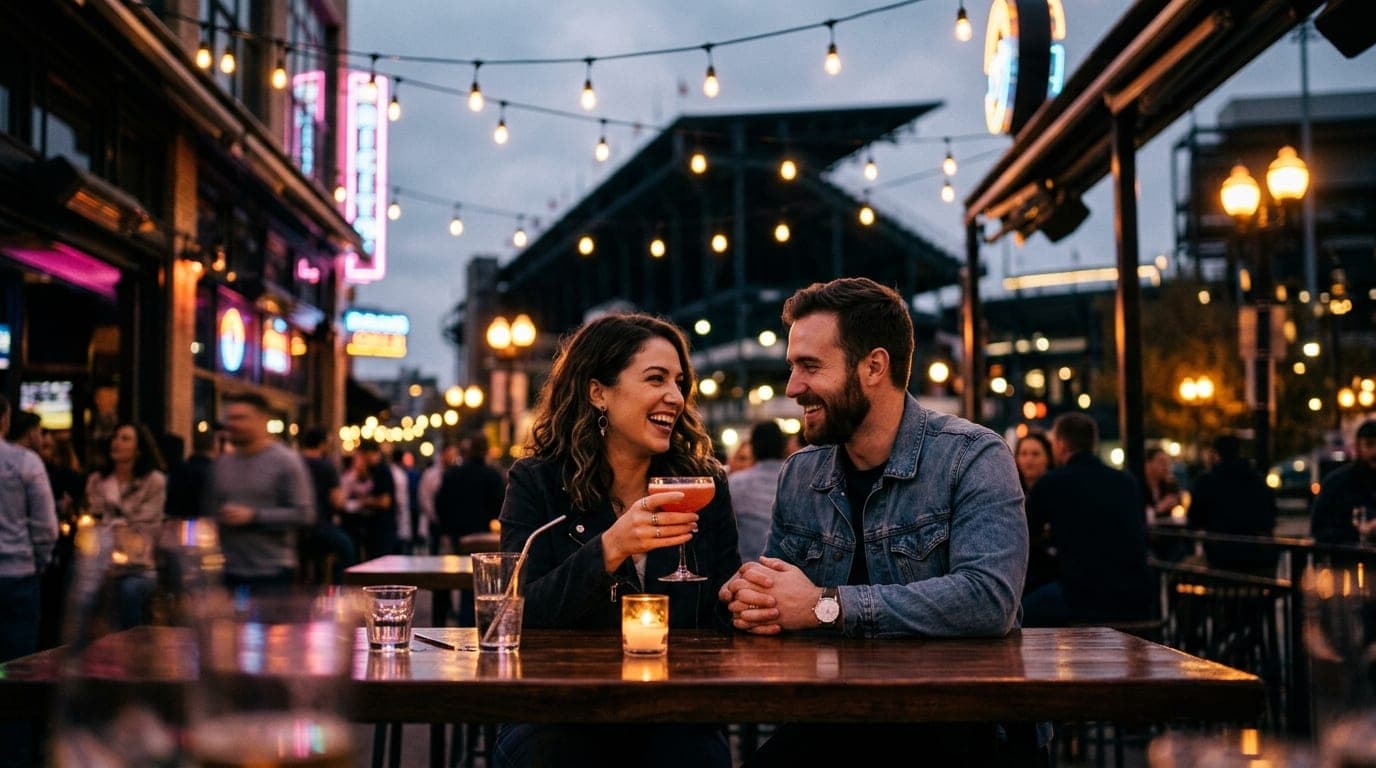 A low-angle, wide-lens shot captures a couple sharing a cocktail at a polished outdoor bar table. Glowing string lights sh...