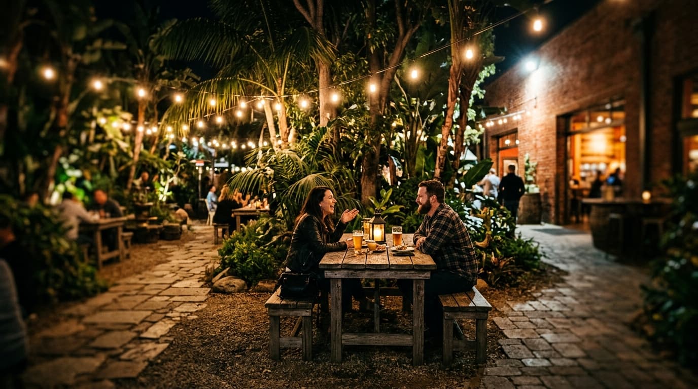 Wide establishing shot, tilt-shift selective focus. A couple sits at a wooden table in Stone Brewing’s garden at night, su...