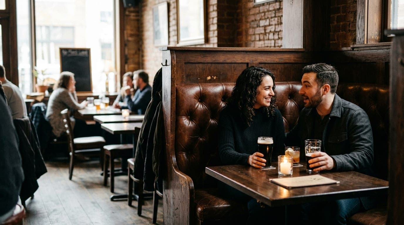 A medium shot captures two people sitting in a deep leather booth at No Man’s Land. Candlelight flickers against dark wood...