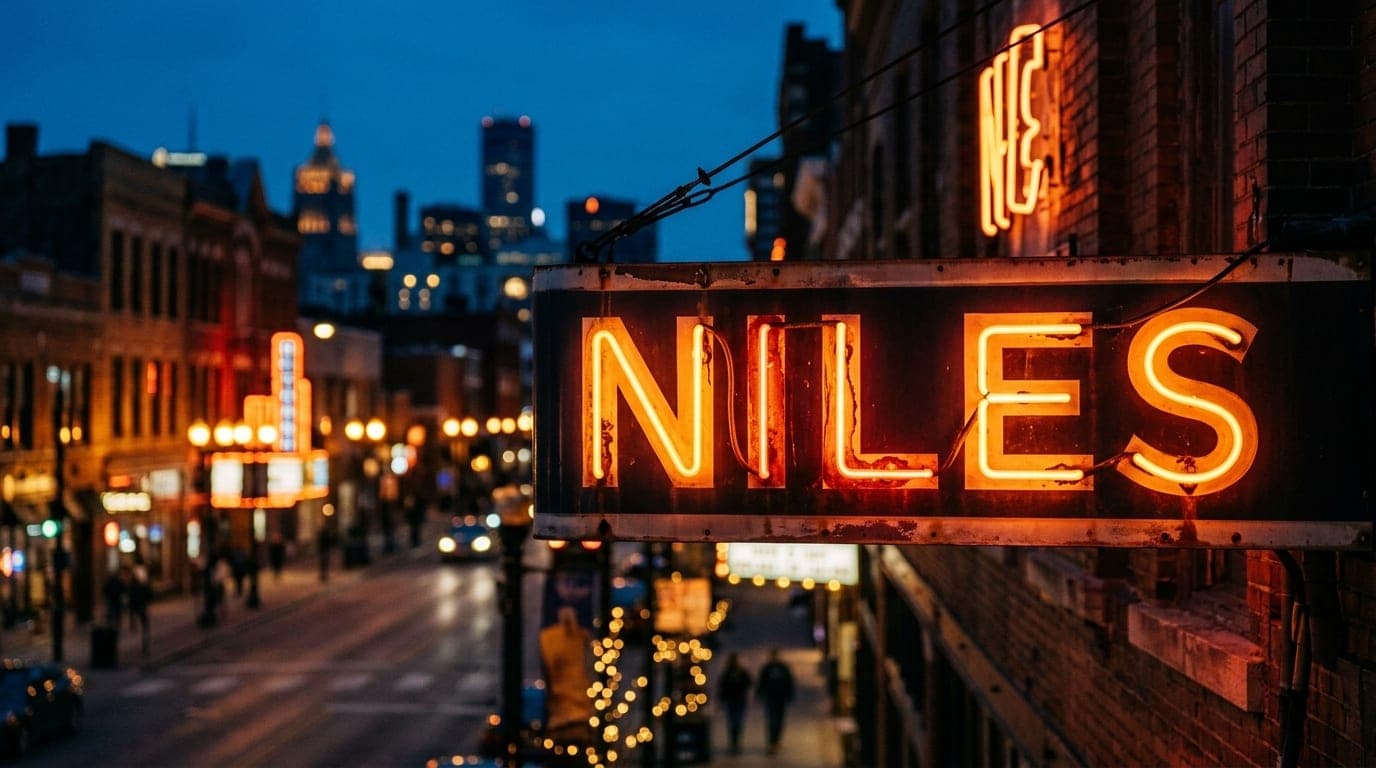 A tight, 100mm macro shot captures the warm, glowing neon sign of a historic Niles storefront at dusk. The blurred, amber-...