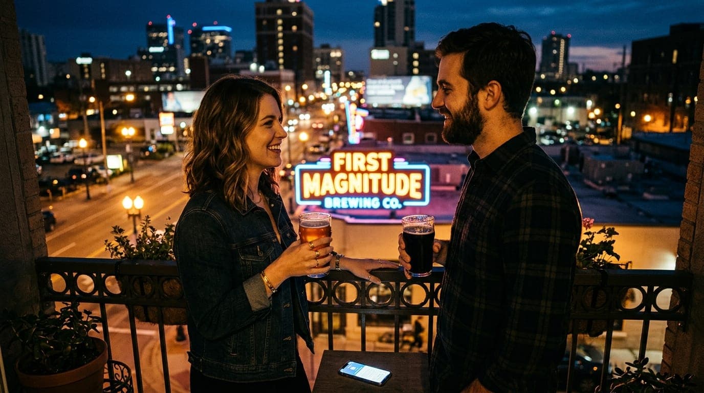 A couple stands on a dark balcony overlooking the glowing First Magnitude Brewing neon sign, holding craft beer pints. A d...