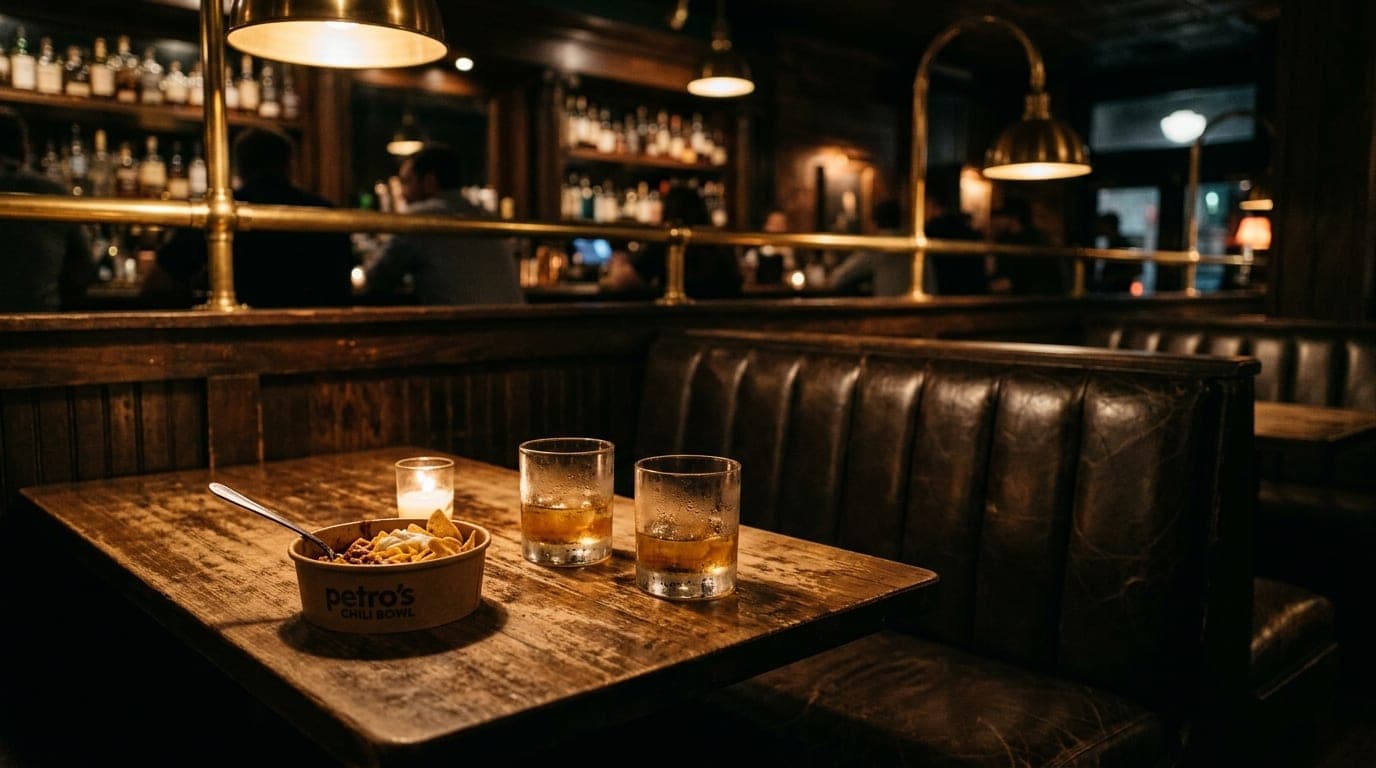 A wide, shallow-depth shot of a dim Knoxville cocktail bar booth. A half-eaten Petro’s chili bowl sits beside two sweating...