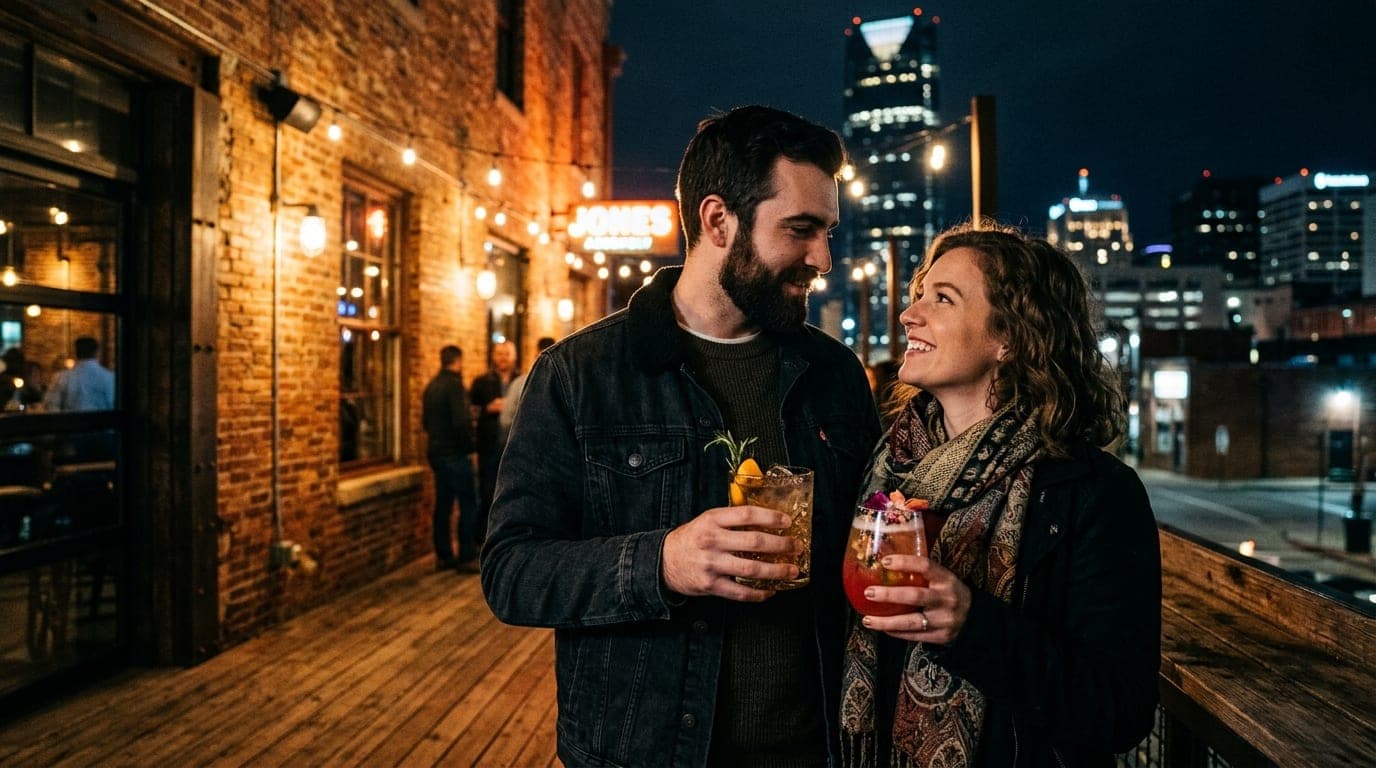 A couple stands closely on the dimly lit outdoor terrace of The Jones Assembly, holding ornate cocktails. The low-angle sh...