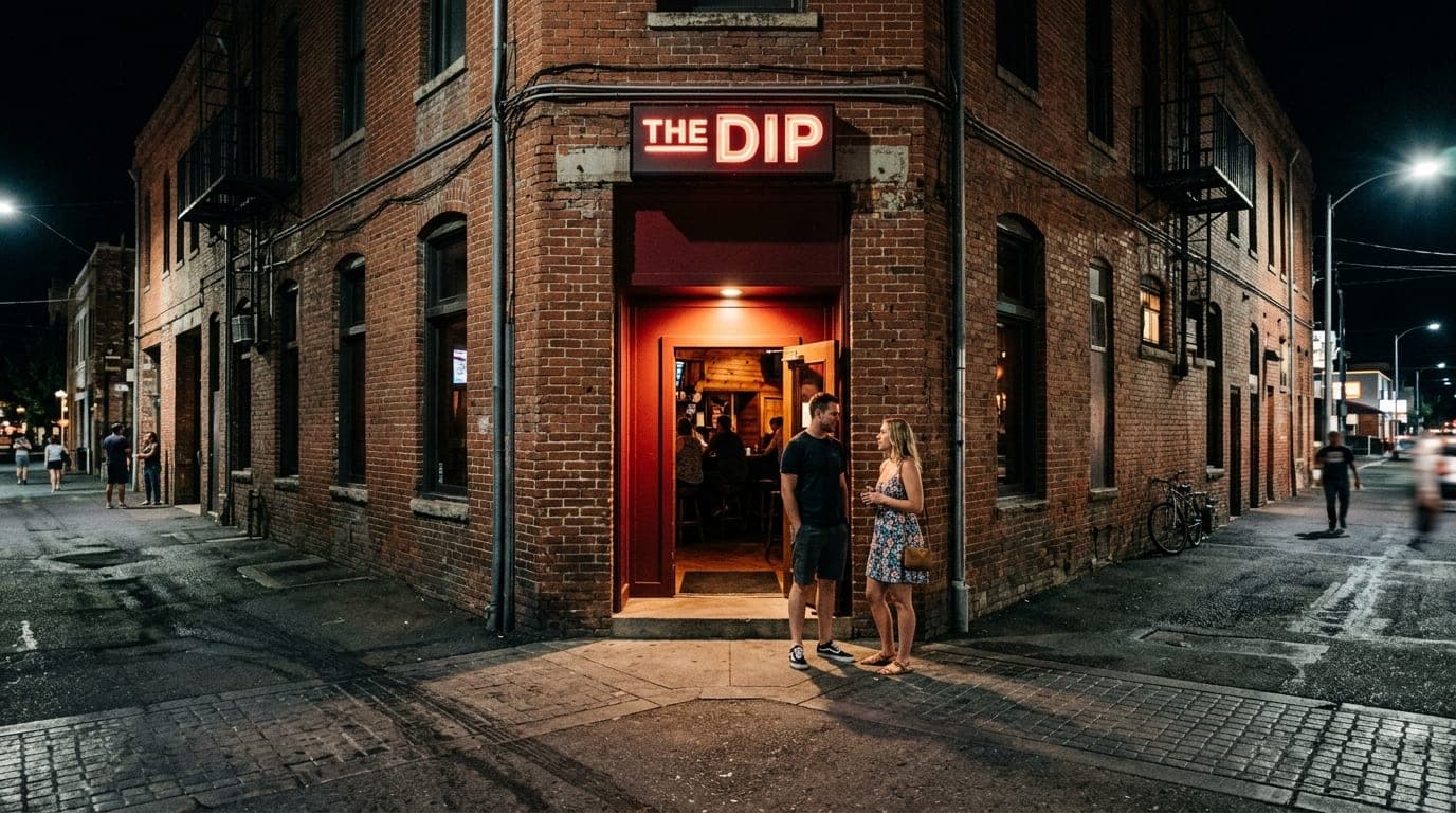 A wide-angle, symmetric shot of The Dip’s entrance at night. A couple in casual summer attire stands near the glowing, dar...