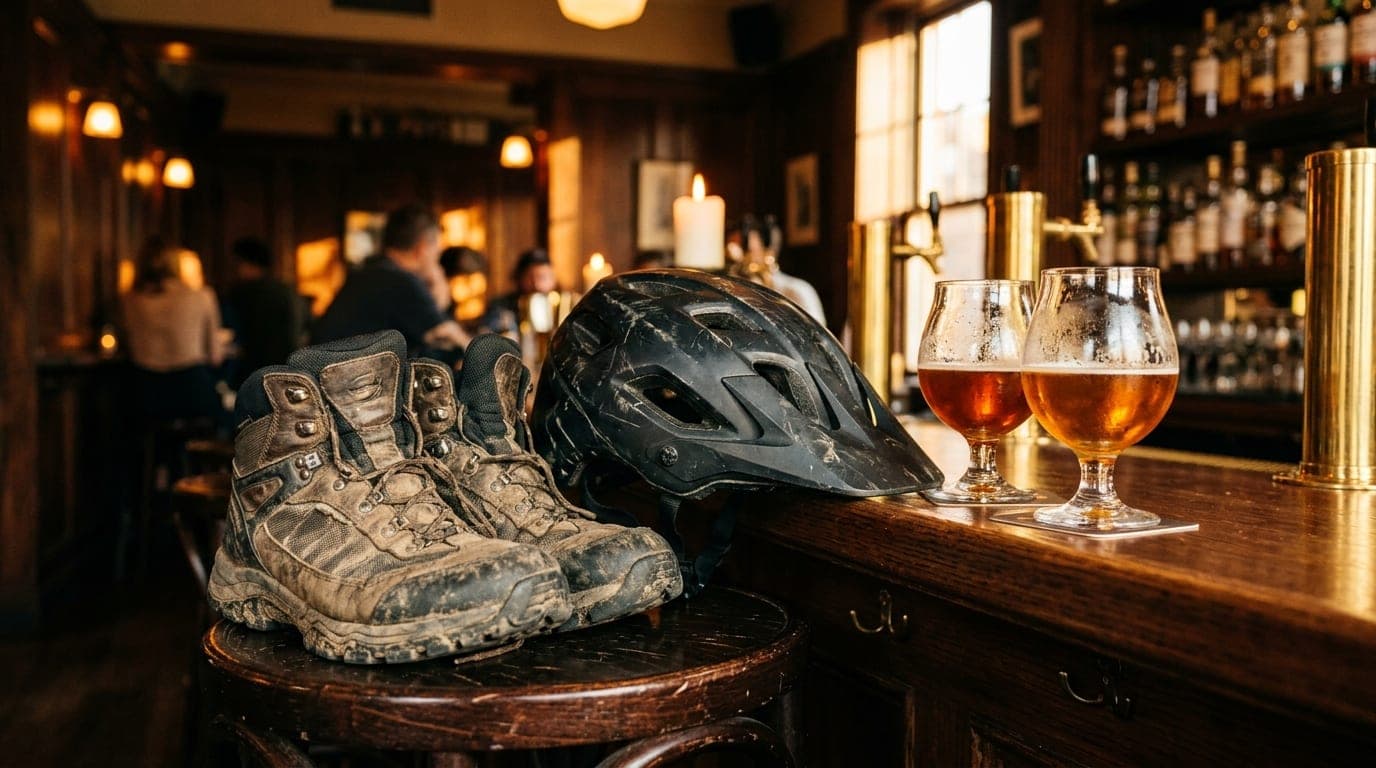 A weathered mountain bike helmet and a pair of worn hiking boots rest on a dark, polished Midtown bar stool beside two amb...