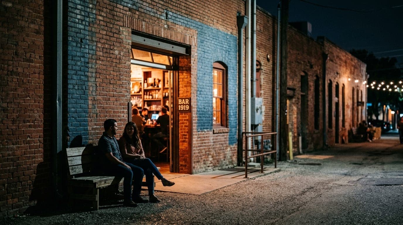 Wide establishing shot of a quiet Blue Star brick warehouse alley at night. A couple sits on a wooden bench, silhouettes f...