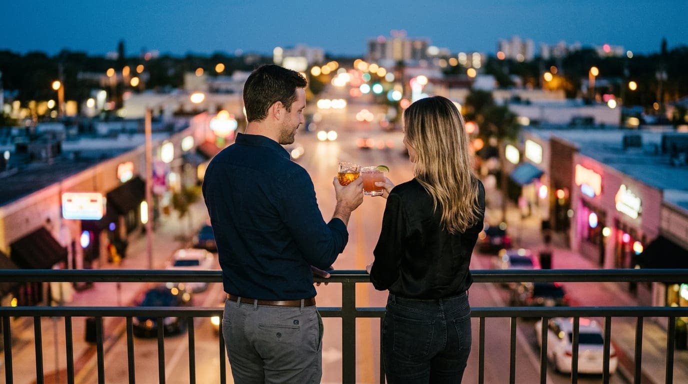 A man and a woman in casual evening attire lean against a balcony railing at Gulf Gate, overlooking blurred, glowing night...