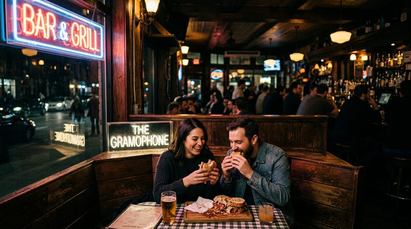 A couple shares a oversized sandwich at a corner booth in The Gramophone, bathed in glowing neon signage. The blurred, moo...