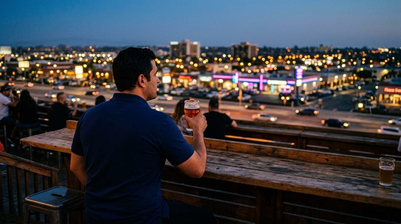 An over-the-shoulder view of a man in a crisp navy polo sitting at a wooden brewery table, glass of craft beer in hand, ov...