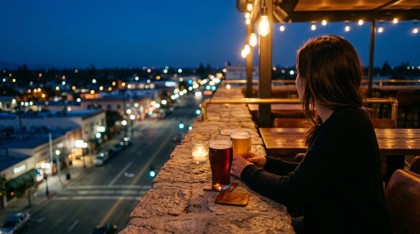 An over-the-shoulder 35mm shot at dusk from a rooftop bar overlooking Main Street. Two glasses of craft beer sit on a ston...